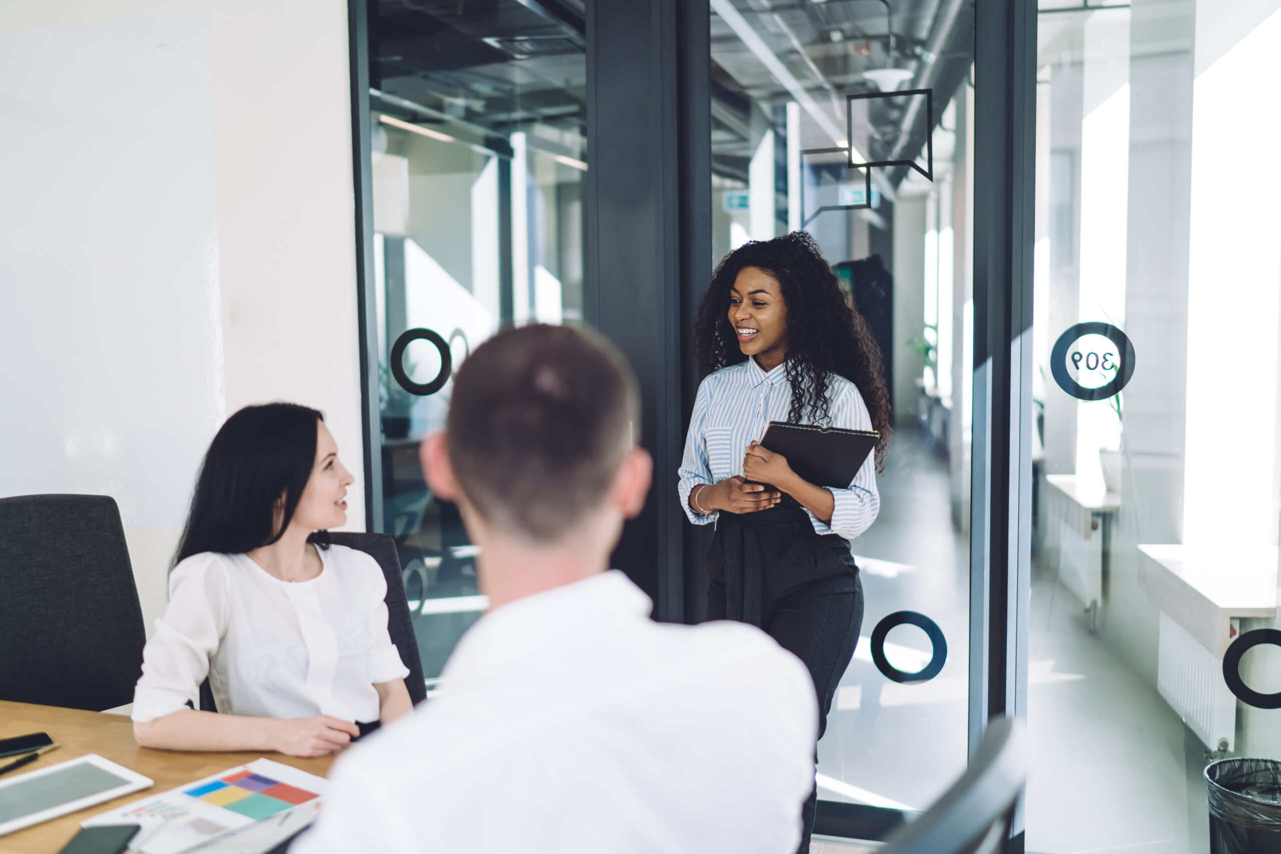 Black curly smiling lady with folder coming in startup company for meeting with hr service about job in modern conference room